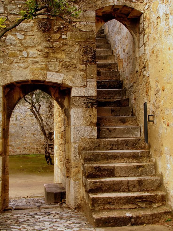 an image of a stone staircase ascending alongside a stone wall, with an archway leading to an outdoor courtyard. the architecture appears old and medieval, with weathered stone and cobblestone paving the ground.