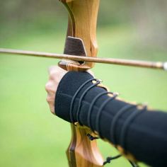 the image captures a closeup view of an archer's hand drawing back a bowstring, ready to release an arrow. the archer's hand is adorned with a protective black armguard.  the blurred green background suggests an outdoor setting, likely a field or forest. the focus on the archer's hand and the tension in the bowstring create a sense of anticipation and highlight the precision required in archery.