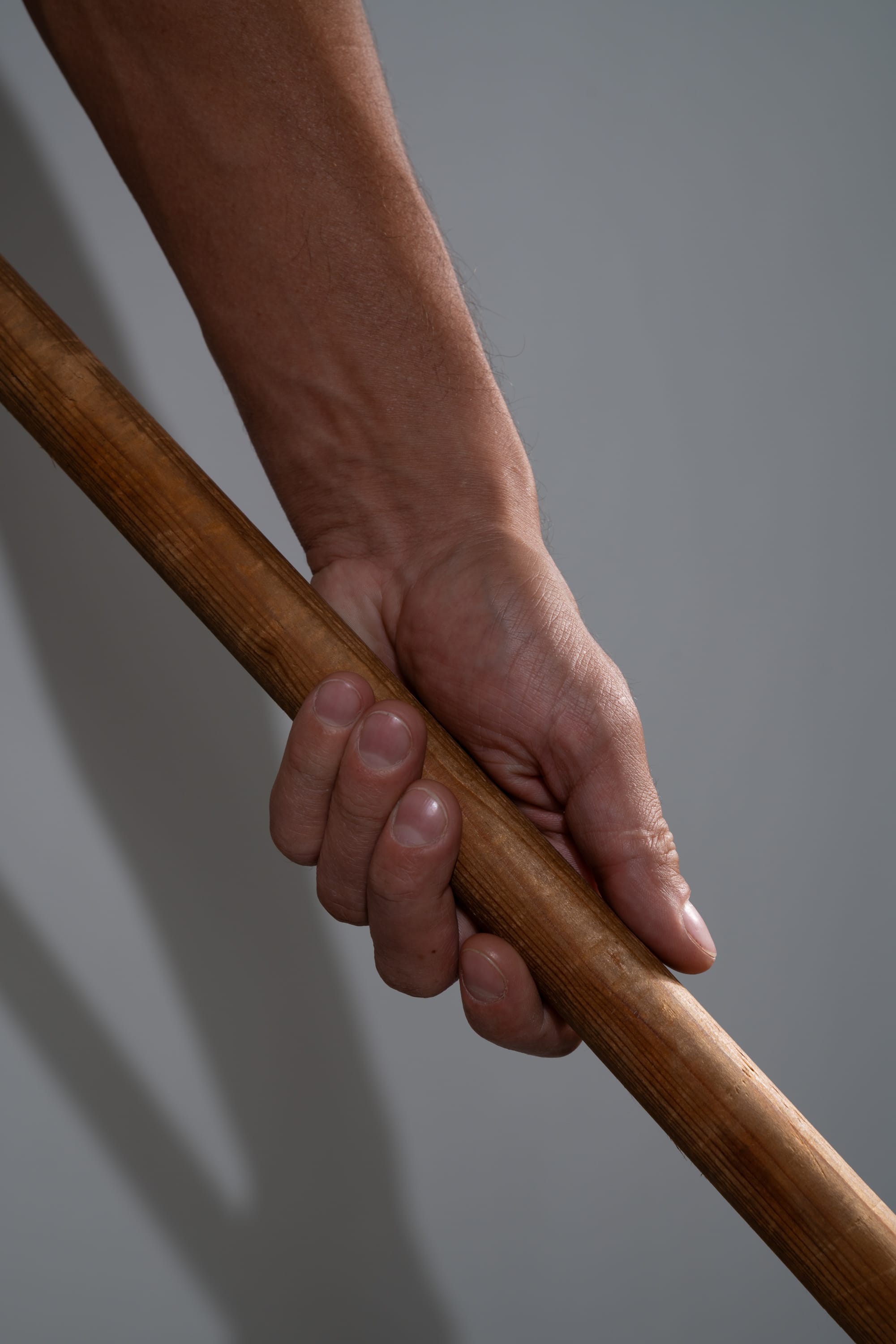 a closeup shot of a man's hand tightly gripping a long, brown wooden stick. the image highlights the texture of both the hand and the wood, with the studio lighting casting a distinct shadow against a plain gray background.