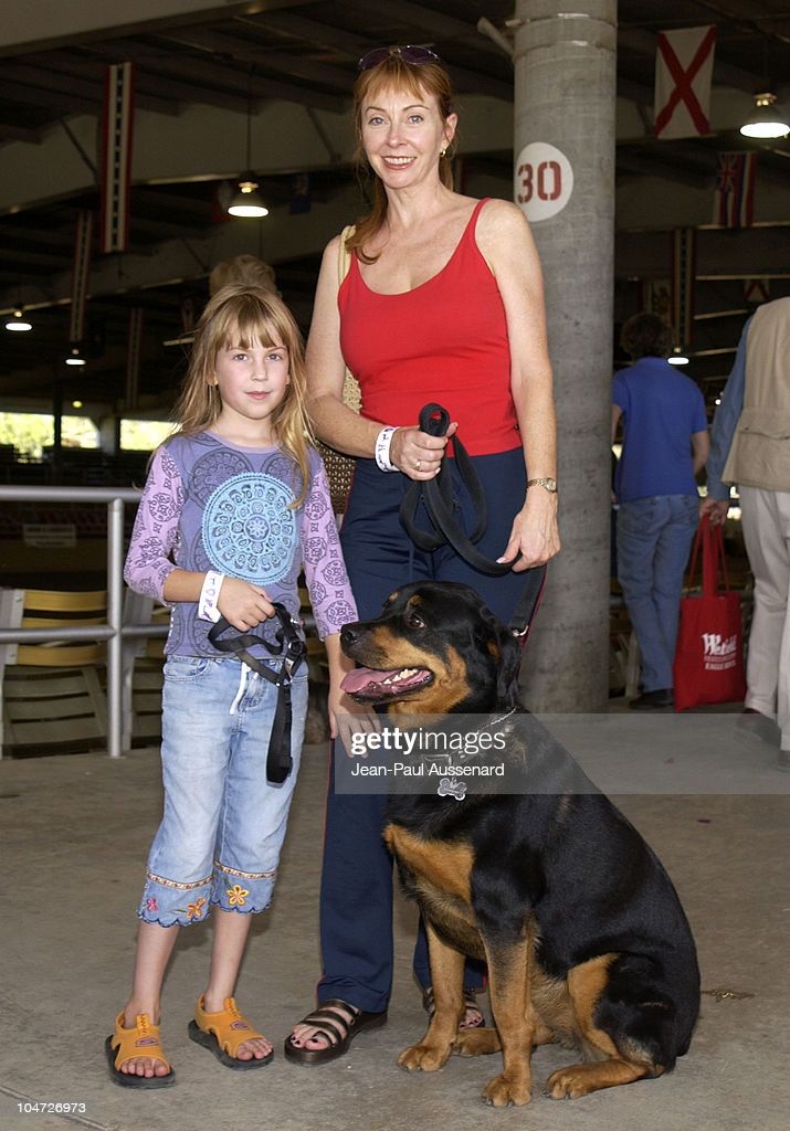 a woman and a young girl are seen outside with a rottweiler. the girl is wearing a pink shirt and blue jeans, and the woman is wearing a red tank top and blue jeans. the dog is sitting between the woman and the girl and all three are smiling at the camera. they appear to be a family, and are out for a day of fun in the sun.