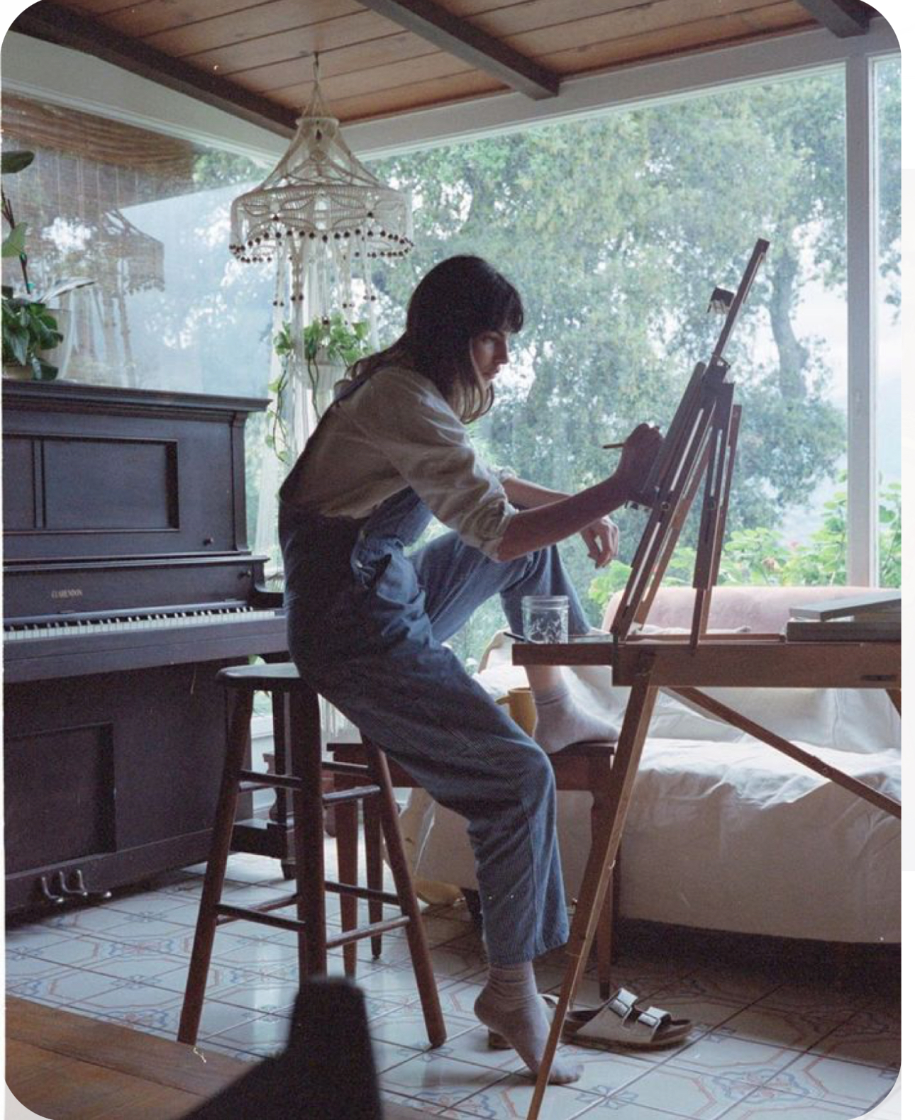 a woman wearing overalls sits on a stool in front of an easel with a paintbrush in her hand as she paints in a naturally lit room. there is a piano, houseplants, and a window in the background.