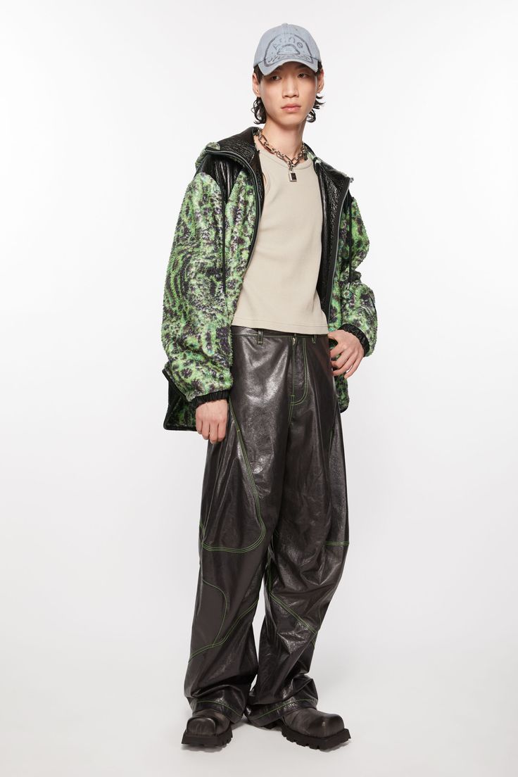 fullbody shot of a male model showcasing a stylish outfit. he's wearing a lime green and black patterned jacket with a black leather hood, a beige crewneck shirt, dark brown leather pants with unique stitching details, and black chunky boots. the model also sports a light grey baseball cap and a simple necklace. the backdrop is a plain white studio setting. the overall style is contemporary and casual, with a blend of streetwear and highfashion elements.  the image is welllit and professionally captured.