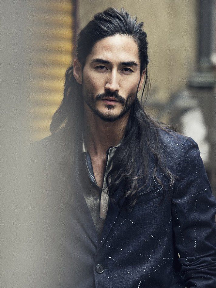 a head and shoulder portrait of a handsome asian man with long, dark hair and a beard, wearing a dark blazer over a collared shirt. he is looking directly at the camera with a serious expression.