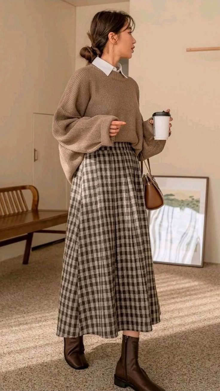 a woman stands in a tan sweater, plaid skirt, and brown boots. she is holding a coffee cup and a brown shoulder bag.
