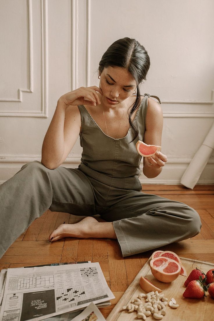 a woman wearing a green jumpsuit is sitting on a wooden floor with her legs crossed. she is holding a slice of grapefruit in her hand. there is a newspaper with a crossword puzzle in front of her and a cutting board with strawberries, cashews, and a grapefruit on the floor beside her. the image is soft and natural, with a relaxed and comfortable vibe. the woman looks relaxed and at ease, as if she is enjoying a moment of quiet contemplation.  the overall image is one of calm and simplicity.