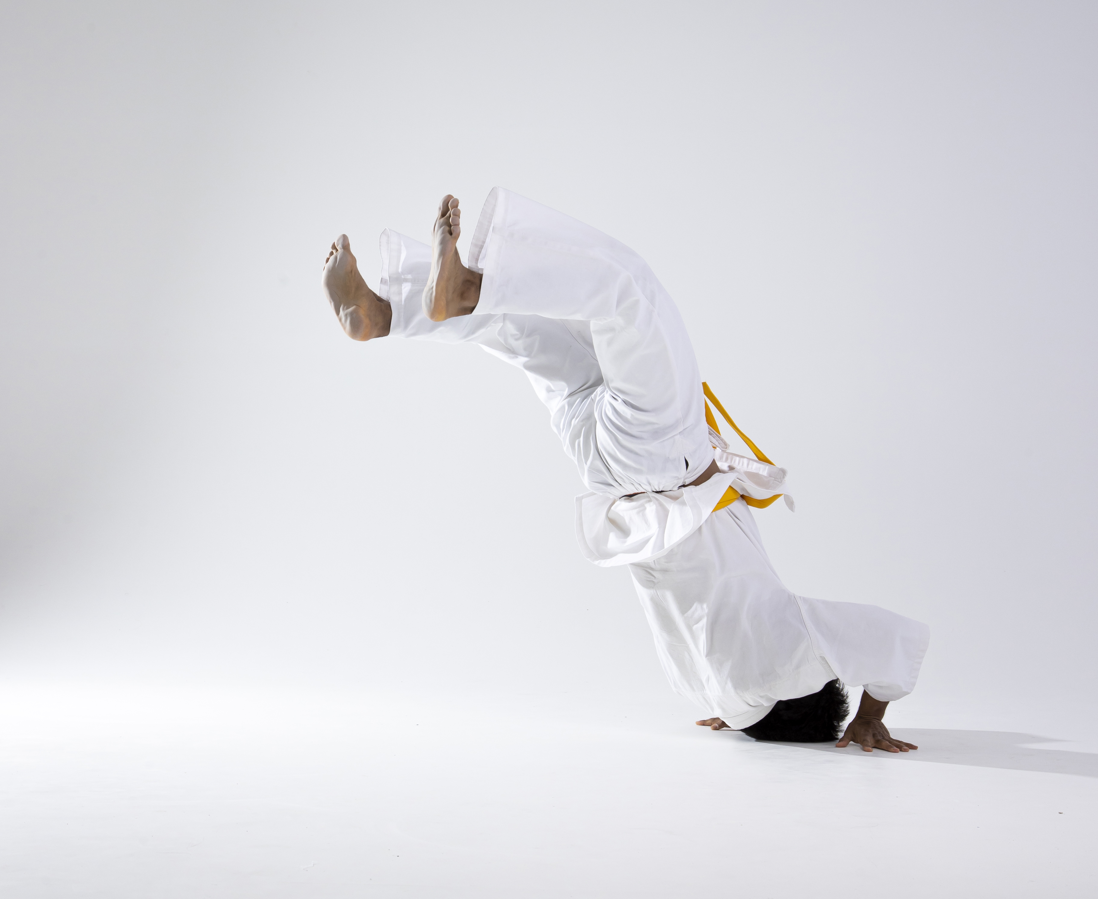 a man in a white martial arts uniform with a yellow belt is performing a headstand. he is upside down with his feet in the air and his head and hands on the ground. the background is white.