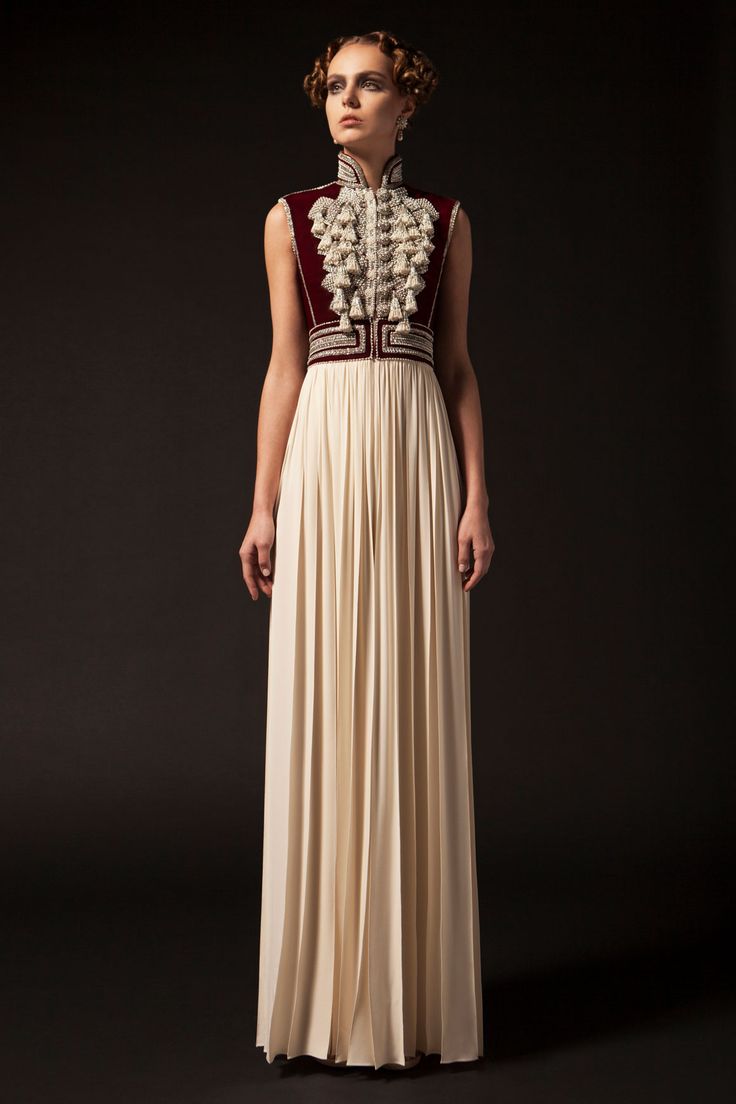 a fulllength shot of a woman in a formal dress, standing against a dark background. the dress features a sleeveless, burgundy velvet bodice with intricate white tassel detailing and a high, ruffled neck. the skirt is a flowing, pleated ivory color. the woman has her hair styled up and wears minimal jewelry.