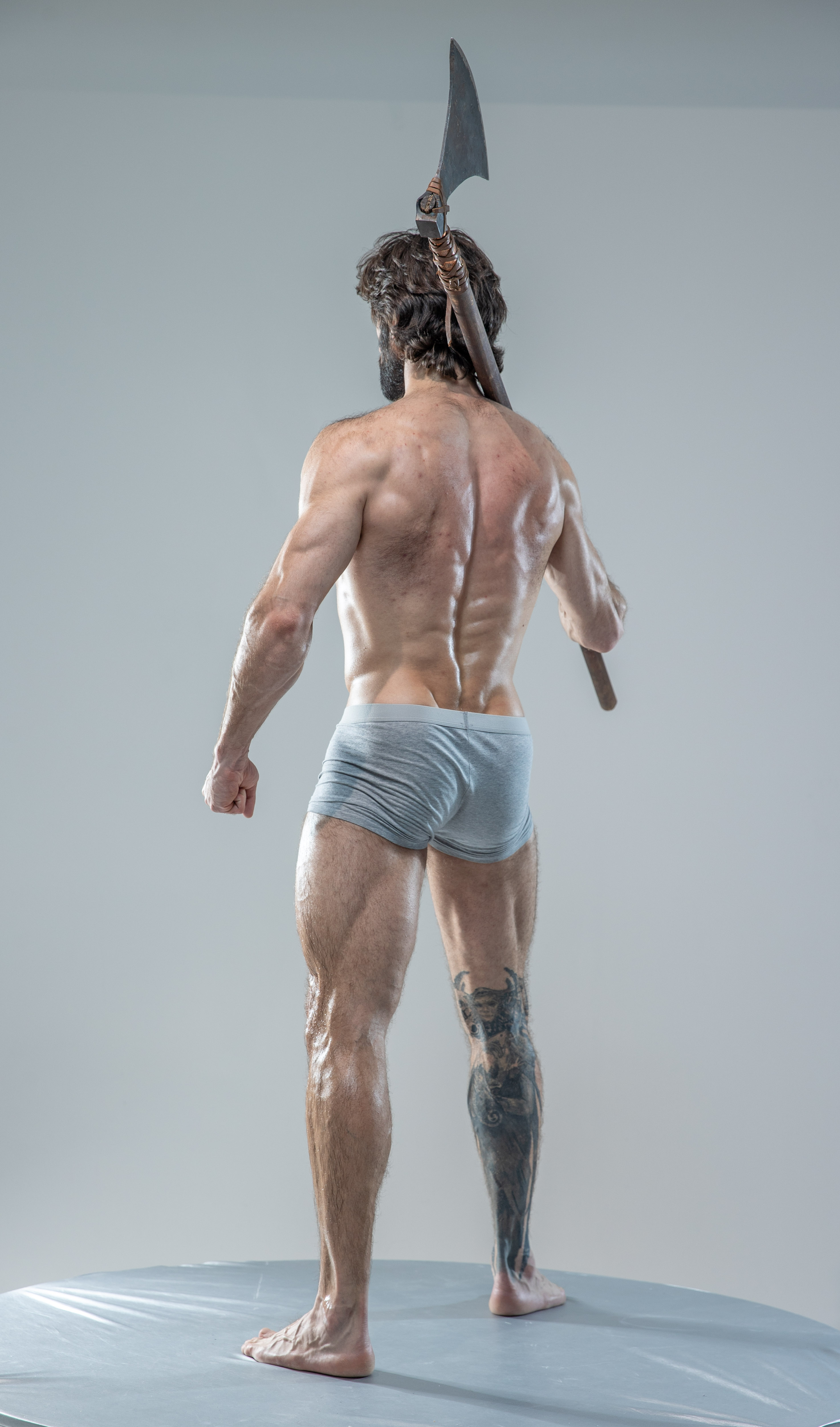 a muscular man with long hair and beard is standing on a platform, facing away from the camera. he is wearing only underwear and holding an axe over his shoulder. the image is a studio shot with soft lighting.