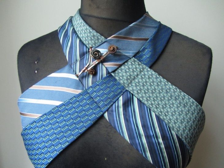 a mannequin is adorned with a unique neckwear piece crafted from several neckties, arranged in a crisscross fashion over the chest area. the ties are a mix of blue and brown colors, with various striped and textured patterns. a pin with a spiral design embellishes the intersection point of the ties, adding a decorative element to the ensemble.