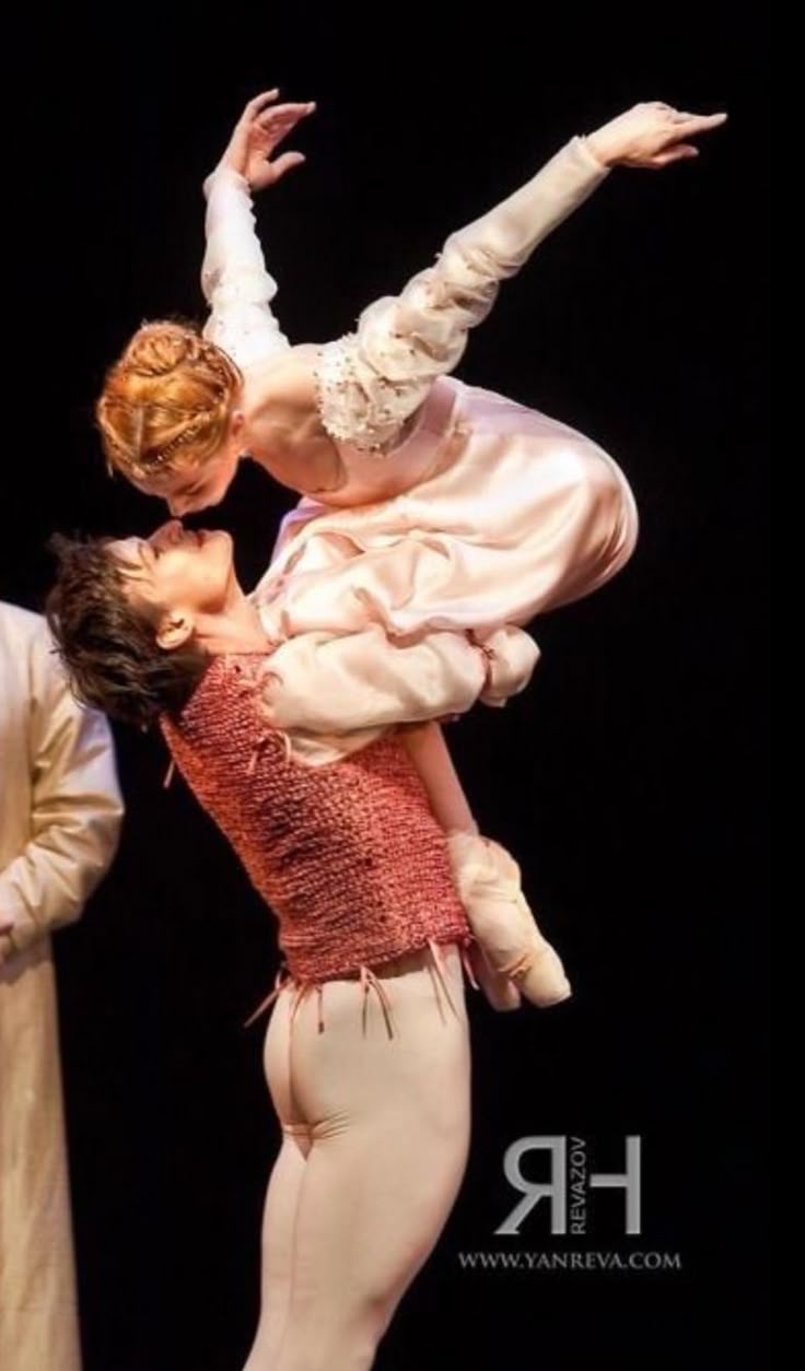 a photo shows two ballet dancers in a close embrace. the male dancer is holding the female dancer in a lift, her face near his. they are in a romantic pose. the background is dark, suggesting a stage setting.