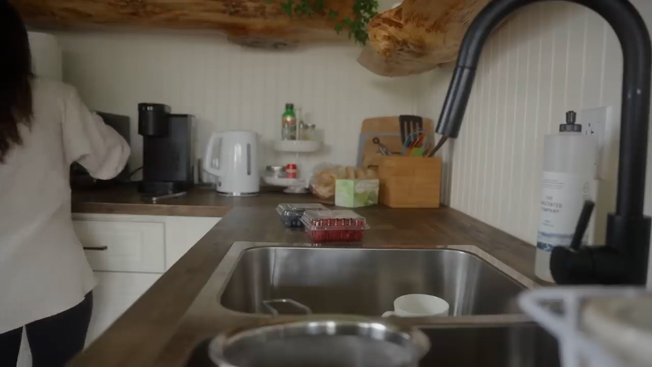 the image shows a person with long black hair wearing a white shirt standing at the counter in a kitchen. in the foreground is a stainless steel sink and a wood countertop.  there are raspberries in a clear plastic container on the counter. the kitchen has white shaker style cabinets and a live edge wood shelf.