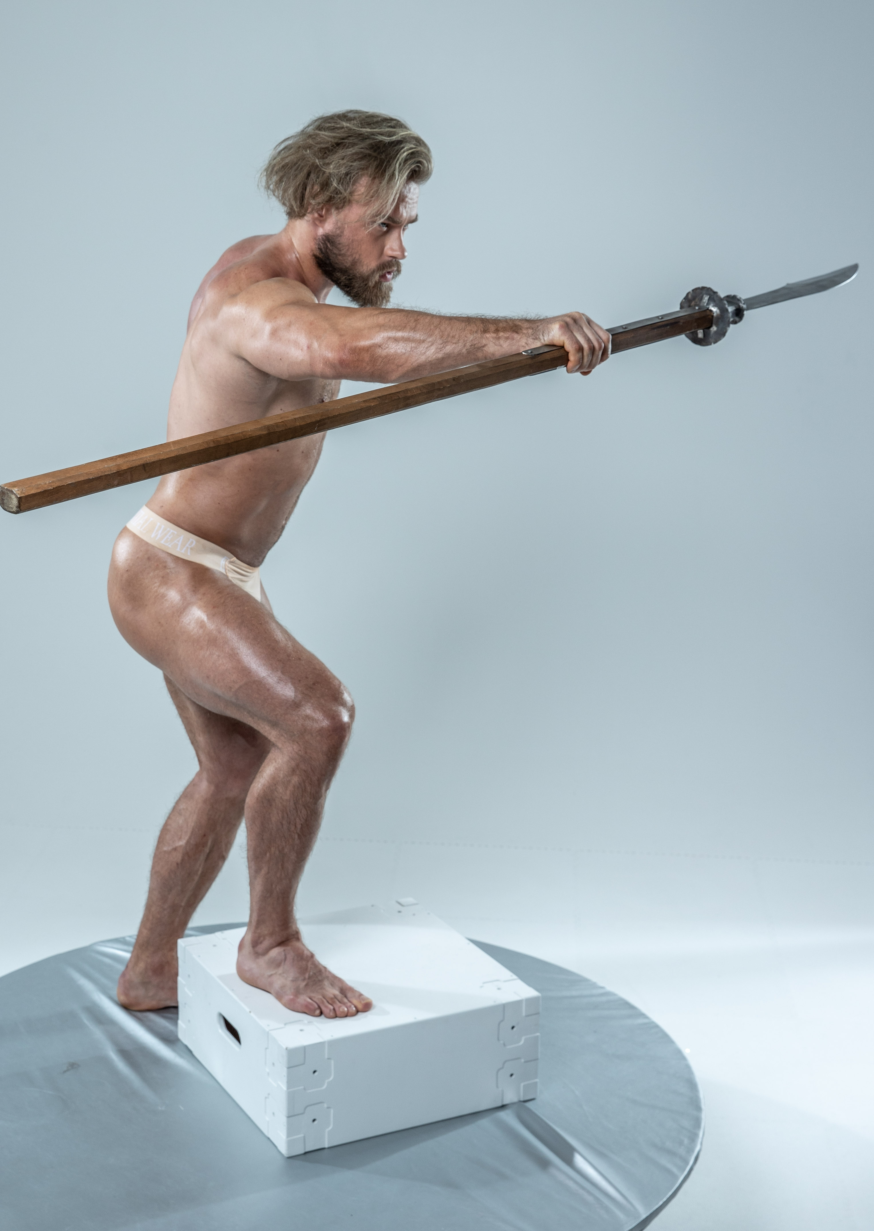muscular man with long blond hair and a beard stands in a dynamic pose on a white platform. he holds a long spear, its wooden shaft grasped firmly in his hands. he wears only a white loincloth, his body tensed with focused energy. the background is a neutral gray, the studio lighting highlighting his physique and the lines of the spear.