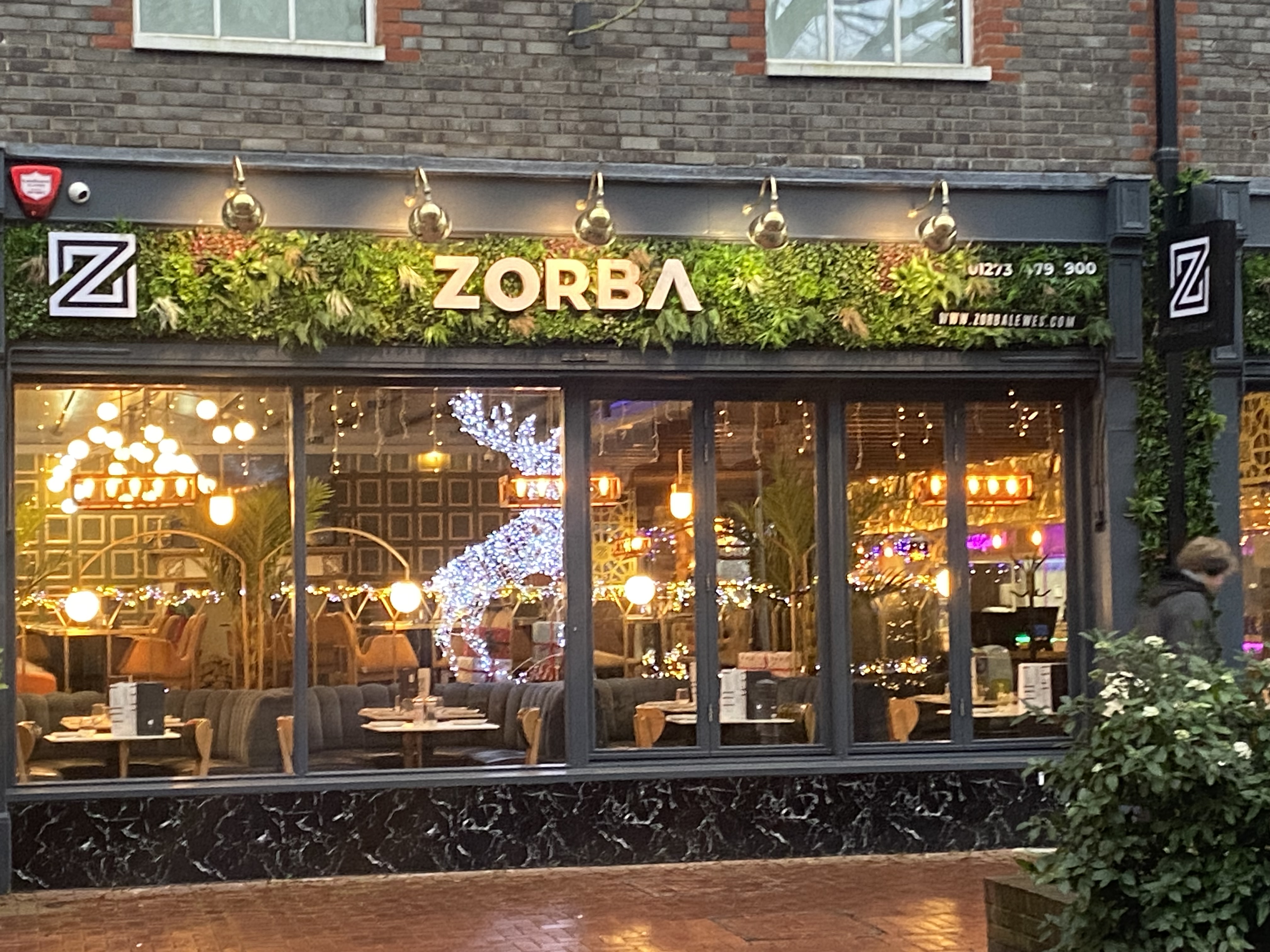 the image shows the exterior of a restaurant called "zorba" in london at night. the restaurant's storefront is modern and stylish, featuring a dark gray facade with a prominent vertical garden above the windows. gold light fixtures and christmas decorations, including a lightup reindeer, are visible inside. the windows reflect the interior dining area, which appears warmly lit and decorated for the festive season.  the overall impression is one of a contemporary and upscale restaurant.  a person is partially visible walking by the restaurant on the right side.