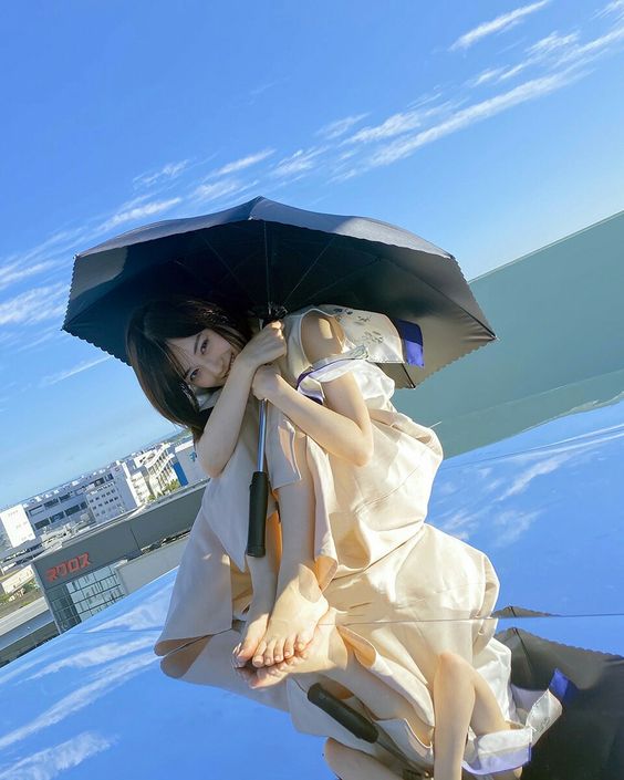 a young woman in a flowing light beige outfit is sitting on a mirrored surface, possibly the roof of a building. she's holding a closed black umbrella and looking directly at the camera with a soft smile. her pose is relaxed, and her bare feet are visible. the mirror reflects her image as well as the surrounding urban environment and the clear blue sky with a few fluffy clouds. the sunlight is bright, indicating a sunny summer day.