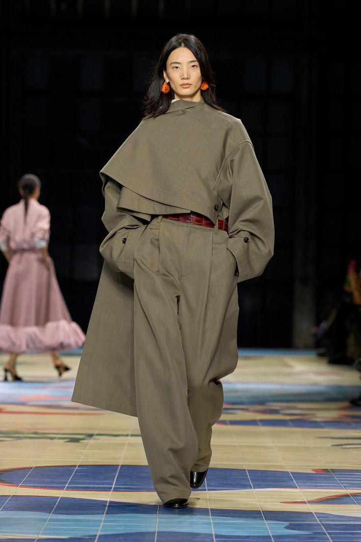 a female model in a brown coat and pants, wearing black shoes and orange earrings, is walking on a runway in a fashion show. the background has a blue, white and brown design on the floor.