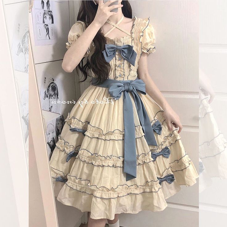 a young woman is taking a mirror selfie in what appears to be her bedroom. she is wearing a creamcolored lolita fashion dress with blue ribbons and bows. the dress is short and has multiple layers of ruffles. she is posing with one hand on her hip. the background of the photo shows a closet with clothes hanging and anime posters on the wall. the lighting is artificial and the perspective is at eye level.
