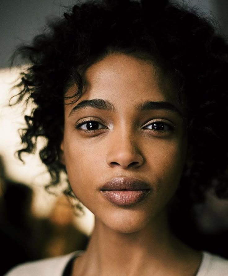 a closeup portrait of a young woman with dark, curly hair and brown skin. she has dark eyes and a neutral expression. the lighting is natural and emphasizes her skin and hair texture.
