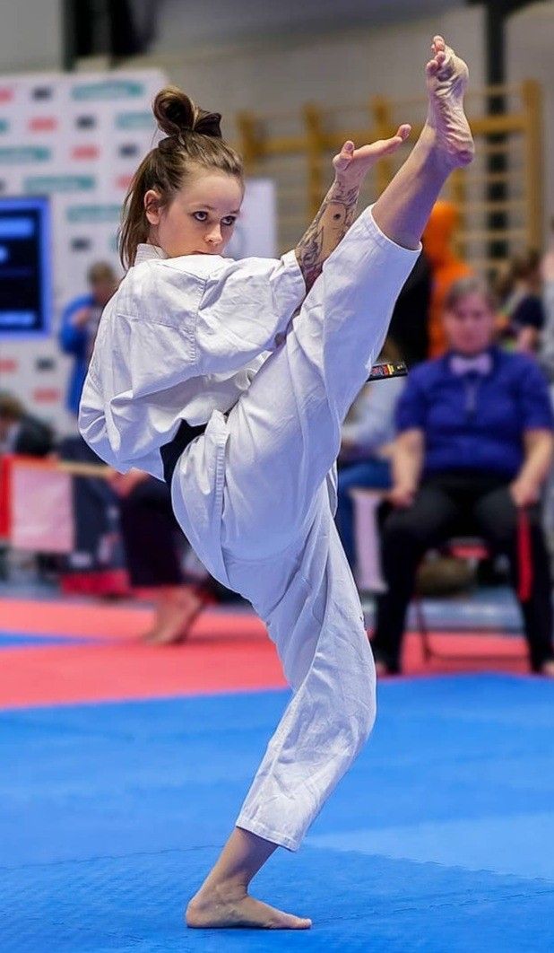 a young woman in a white karate uniform is performing a high kick during a competition or training session. she is focused and determined, showcasing her power, balance, and agility. the background shows a blurred view of other athletes, spectators, and the indoor gym setting.