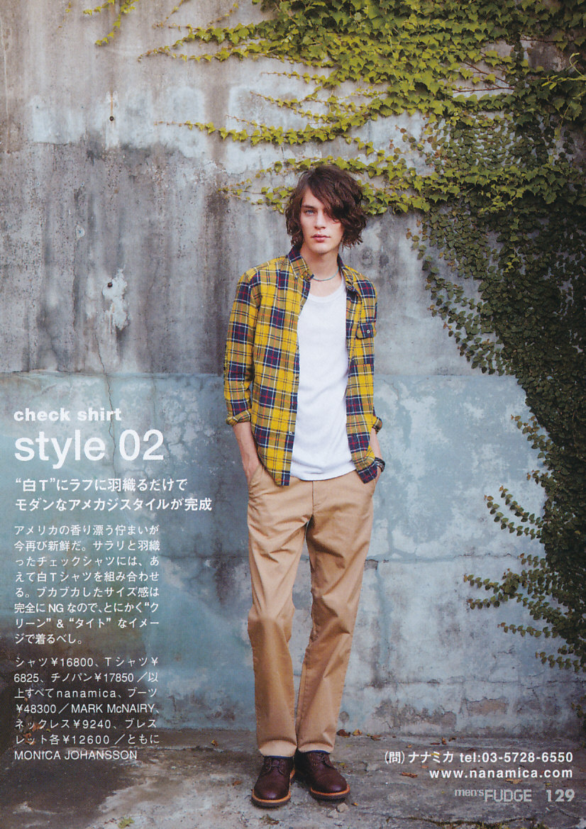 a fulllength image features a male model standing against a textured wall partially covered with vines. he is dressed in a casual yet stylish outfit consisting of a yellow and blue plaid shirt worn open over a plain white tshirt, khakicolored chinos, and brown leather boots. the model has mediumlength brown hair and a relaxed posture. the composition is framed with japanese text and pricing details on the left and contact information on the bottom right, suggesting it is an advertisement from a japanese fashion magazine.
