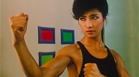 a muscular woman in a black tank top and short hair is striking a martial arts pose with her arm raised and fist clenched. she is in a room with colored squares on the wall, and her expression is focused and determined.  she is likely training or preparing for a fight.