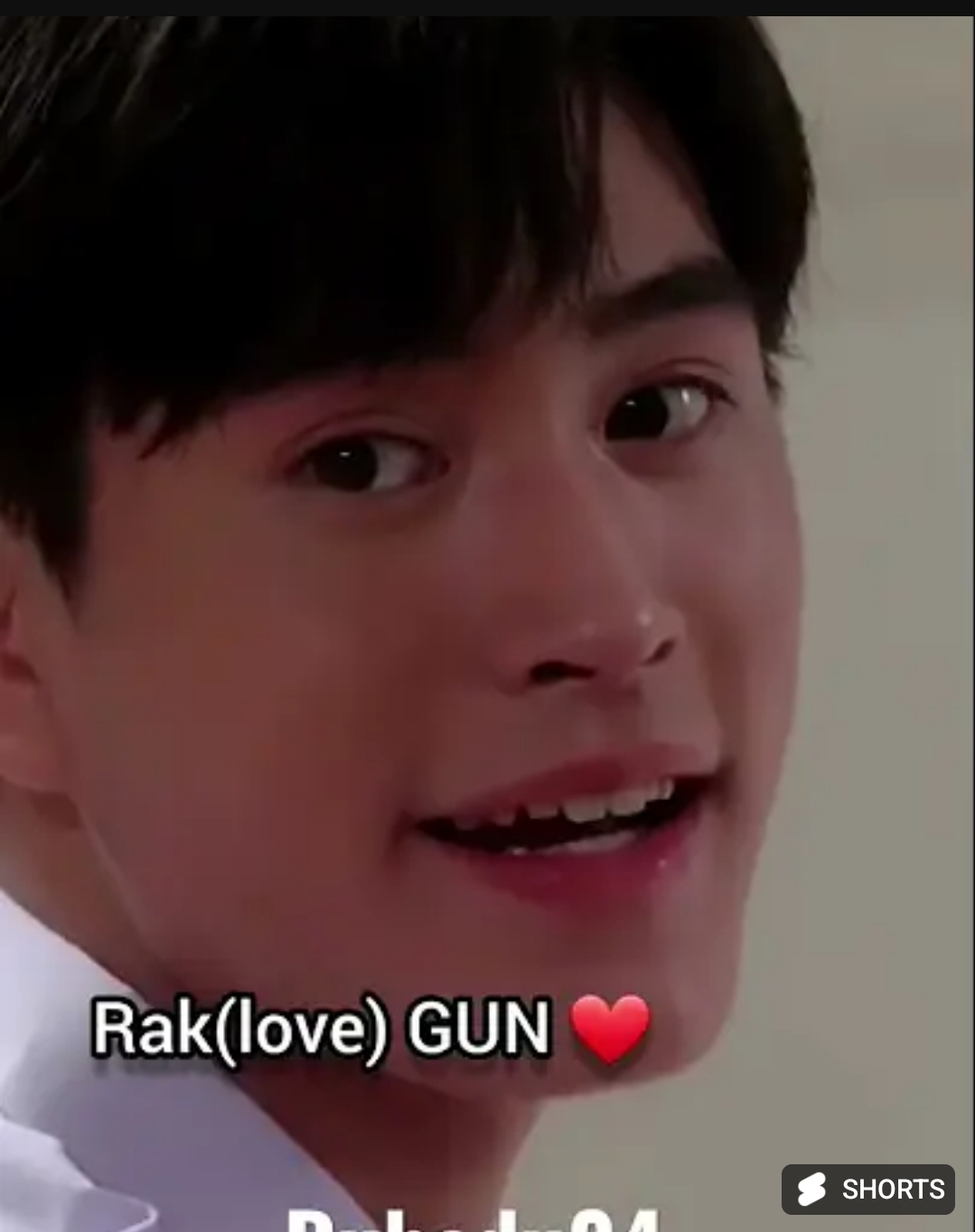 a closeup portrait of a young asian man smiling. he has short black hair and is wearing a white shirt. the lighting is soft and the background is out of focus. the text "rak(love) gun" with a red heart emoji is superimposed on the image.