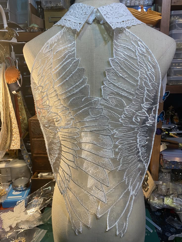 the image shows a white lace collar with attached angel wingshaped lace appliques displayed on a mannequin. the collar has a classic shape with delicate lace trim, while the lace wings are intricate, with detailed featherlike patterns on a sheer fabric. the setting appears to be a design studio, with various craft supplies and sewing equipment visible in the background. the piece is likely a handmade clothing accessory or embellishment for couture fashion.