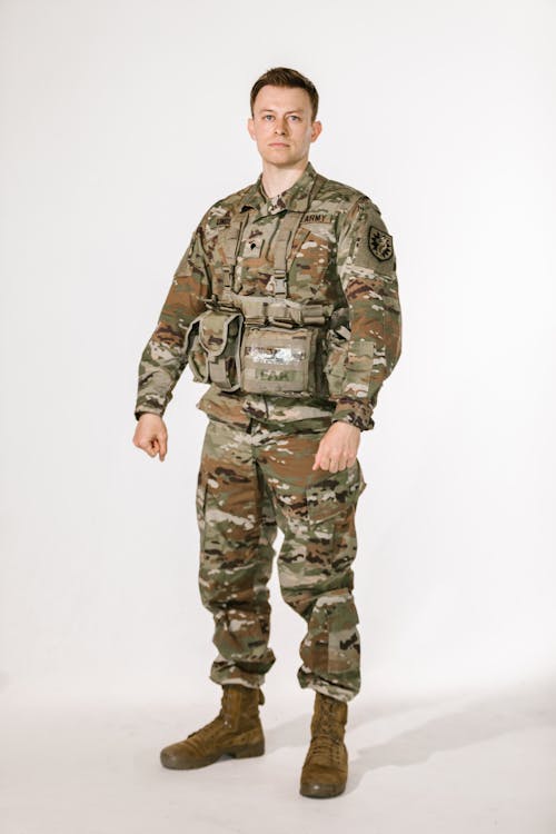 a fullbody studio shot of a male soldier in a u.s. army operational camouflage pattern (ocp) uniform. he is wearing a vest with pouches and is standing against a plain white background. his expression is serious and he appears to be in a formal or official pose. the image is welllit and clearly shows the details of his uniform and equipment.