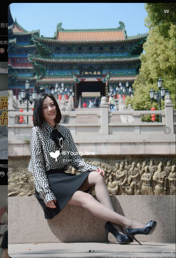 a young woman, dressed in black and white fashion, stands with her legs crossed and smiles at the camera as she sits on a stone wall against a background of traditional chinese architecture.