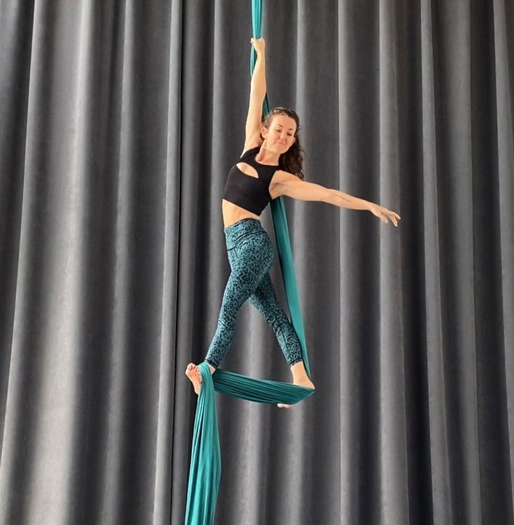 a woman performs an aerial silks routine. she is suspended from a teal silk fabric while doing a pose. she is wearing a black sports top and teal leopard print leggings. the background is a grey curtain.  she is smiling and looking at the camera.