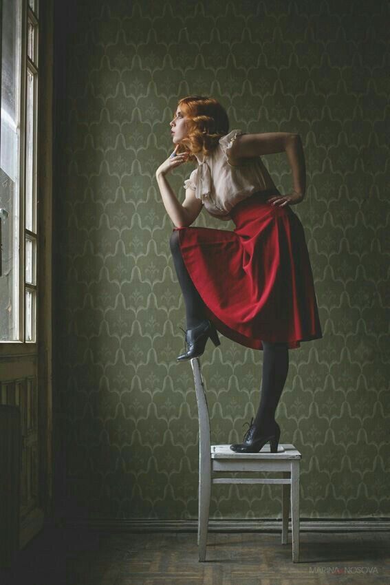 a woman with red hair stands on a white chair, posed near a window in a room with patterned green wallpaper. she wears a beige blouse, a red skirt, black stockings, and heeled boots. her hand is near her chin as she looks out the window. the scene has a vintage or retro aesthetic, and the overall mood is thoughtful and artistic.