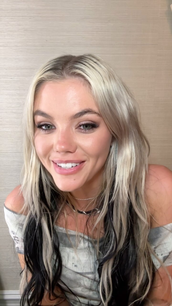 a portrait of a woman with blonde hair and black streaks. she is smiling and wearing an offtheshoulder shirt and a necklace. she also appears to be wearing makeup. the background is a beige wall.