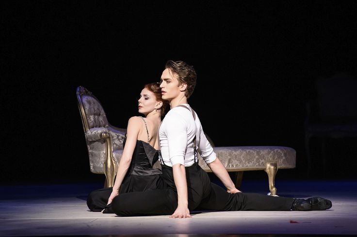 a male and female ballet dancer are performing on stage. the male dancer is in a split position, with his leg stretched out to the right. the female dancer is sitting in front of him, with her back to the audience. they are both dressed in black. there is a chair in the background and a chaise lounge to the right. the stage is dark, but the lighting is highlighting the dancers. the dancers are performing a graceful and elegant dance routine.