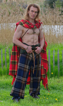 a fullbody shot of a man standing in a grassy field. he is wearing a traditional scottish kilt and plaid, and he is barechested. the man appears to be posing for a photograph.
