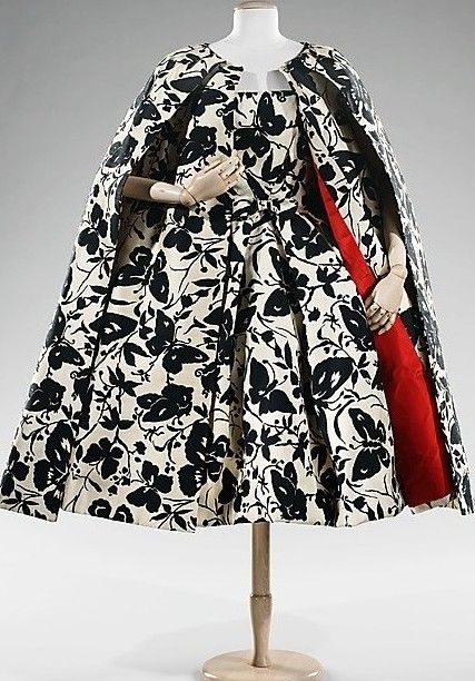 a vintage dress with a cape is displayed on a mannequin. the dress features a black and white floral pattern and a pleated skirt. the cape has a red lining.