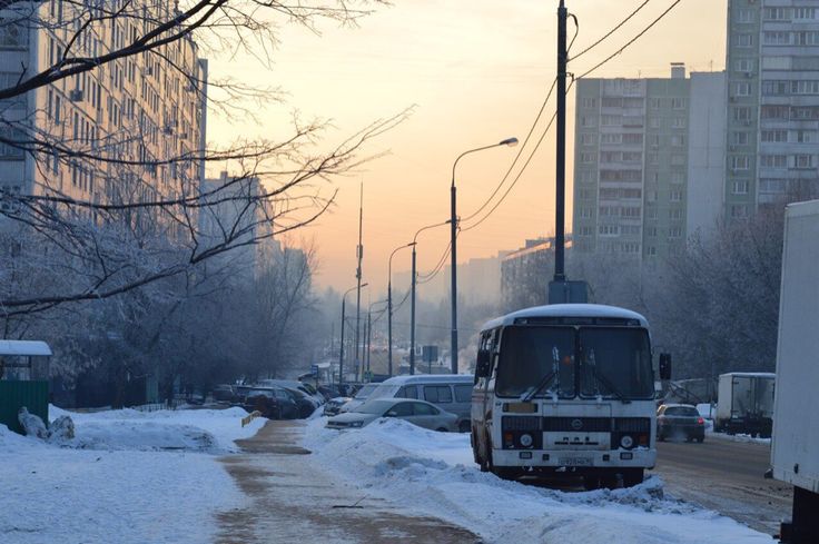 the image depicts a city street scene in winter.  snow covers the ground and sidewalks.  the street is lined with tall, multistory apartment buildings typical of sovietera architecture.  a large, old paz bus is parked at the side of the road, along with several other cars.  bare, snowdusted trees line the street, and streetlights and power lines stretch down the road. the sun is setting or rising, casting a soft, warm light in the background, creating a somewhat melancholic and peaceful atmosphere.  the overall impression is of a cold, quiet, but still somewhat active urban setting in a city, possibly in russia.