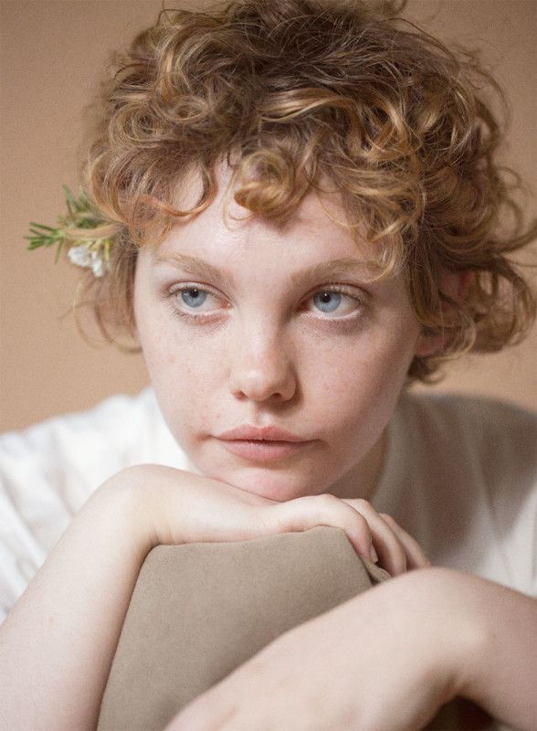 a closeup portrait of a young woman with blonde, curly hair and striking blue eyes. she has fair skin with visible freckles. a small sprig of flowers is tucked behind her ear. she rests her chin on her hands, giving her a pensive expression. the background is a soft beige color, and the lighting is gentle, creating a soft and intimate mood.