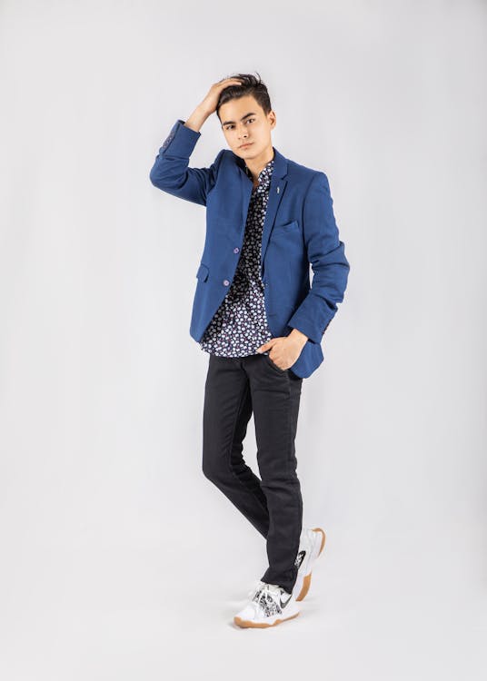 a fullbody studio shot of a teenage boy (or young man) confidently posing against a plain grey background. he's dressed in smart casual attire: a navy blue blazer over a black and white patterned shirt, darkcolored jeans, and white sneakers. he has one hand casually placed in his hair.  the overall style is clean and contemporary.
