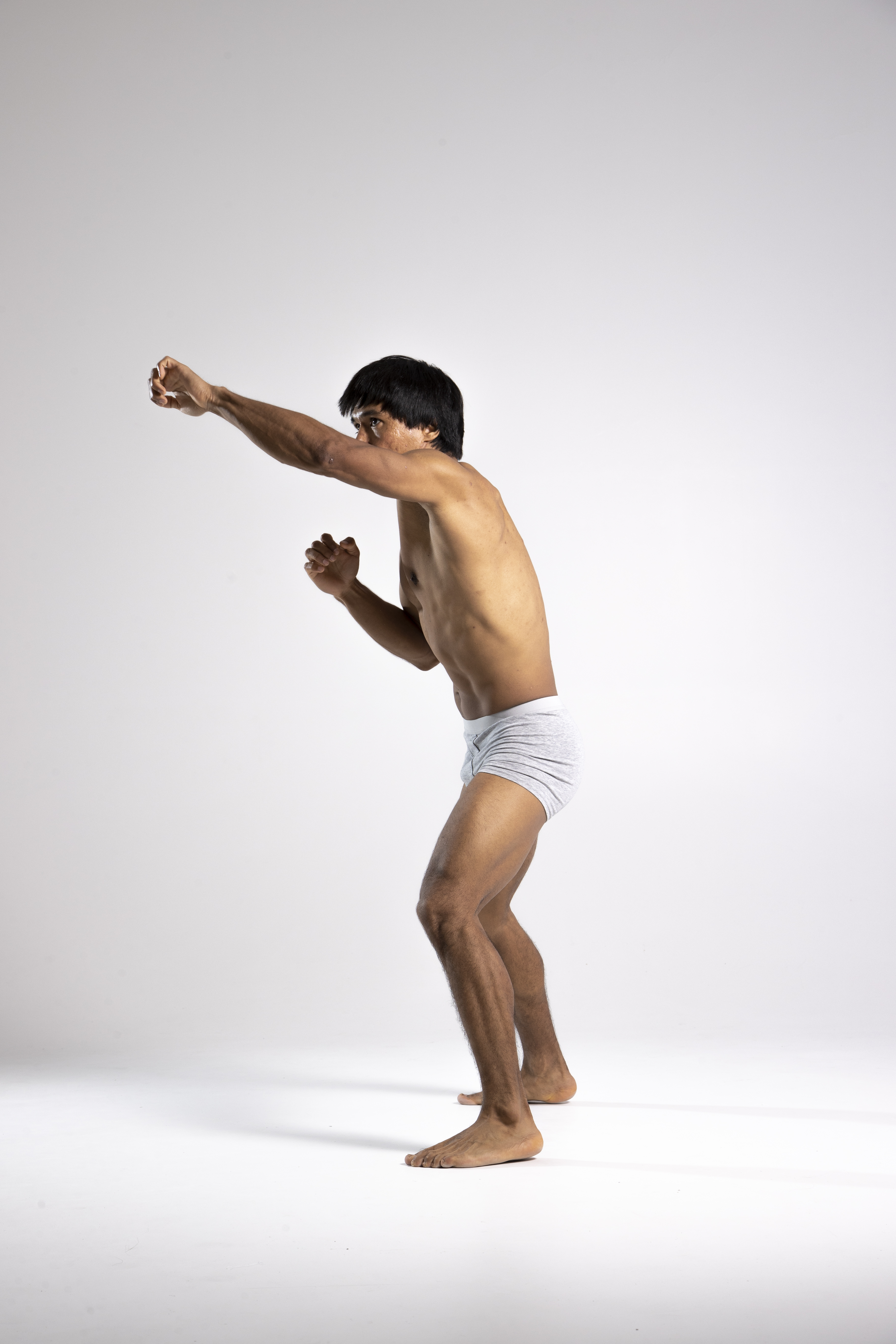 a full body side shot shows a muscular, barefoot asian man in a boxing stance wearing gray underwear. he is determined, with one fist extended in a punch and the other guarding his face. the shot is taken in a studio with a white background.