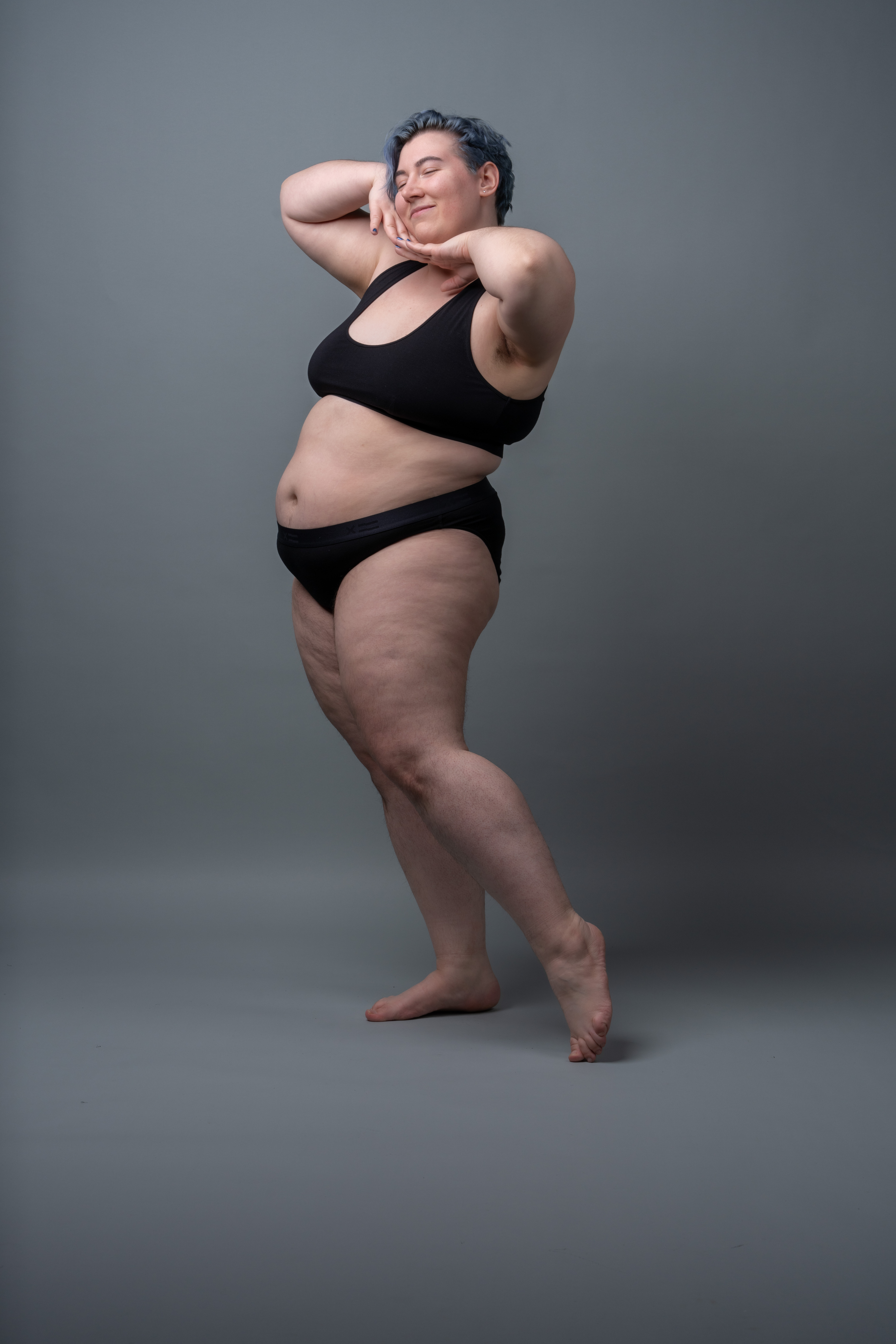 a full body studio shot of a plus size woman with short blue hair, wearing black underwear, smiling with her eyes closed and posing gracefully on a grey background with bare feet. she embodies body positivity and selfacceptance.