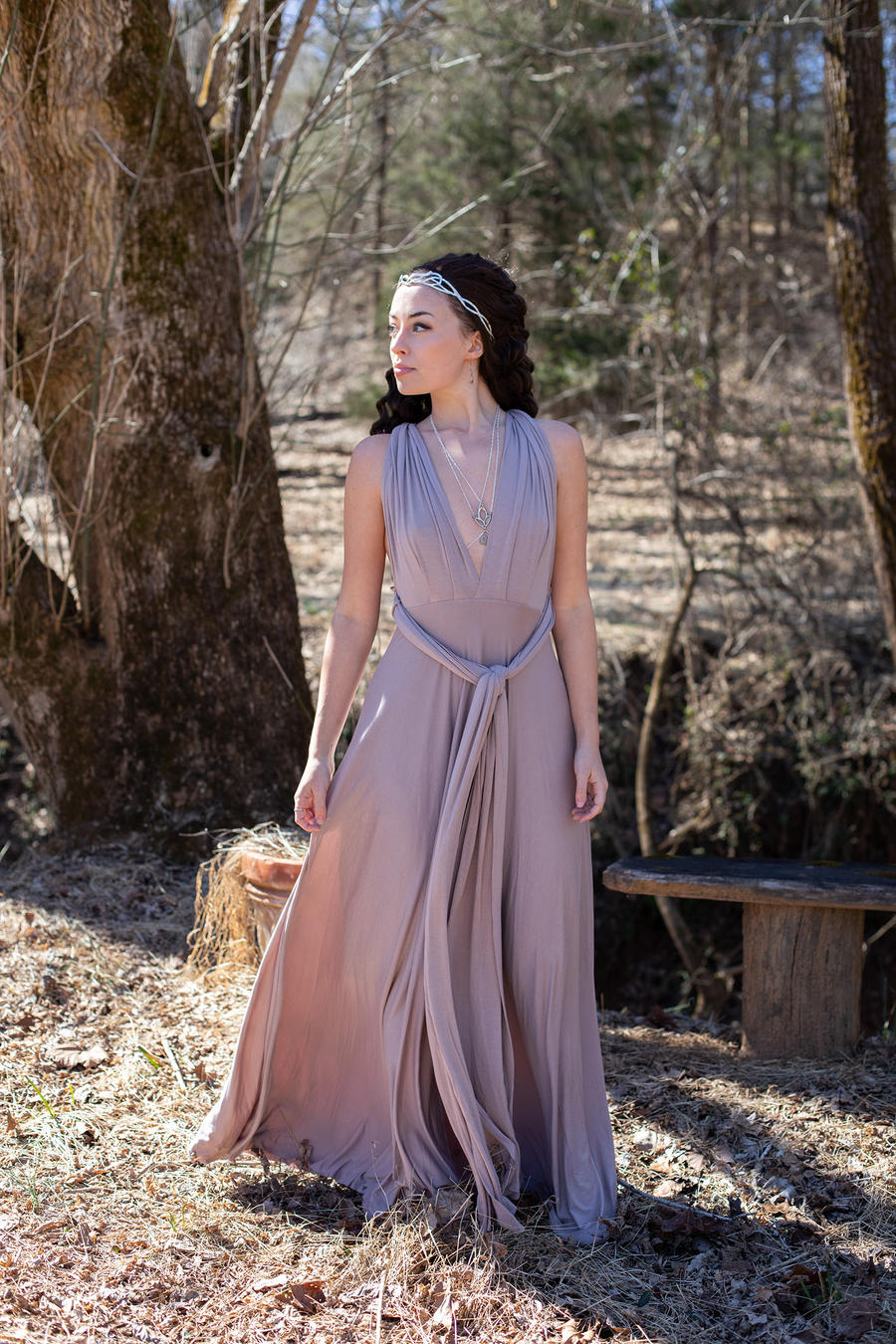 a woman in a long, flowing mauve maxi dress stands in a woodland setting.  the dress is sleeveless and wraps around her waist, tied in a knot at the front. she wears a delicate silver headpiece and several necklaces.  she is looking off into the distance, away from the camera, and appears serene and calm amidst the natural surroundings. a rustic wooden bench is visible in the background. the overall atmosphere is peaceful and elegant.