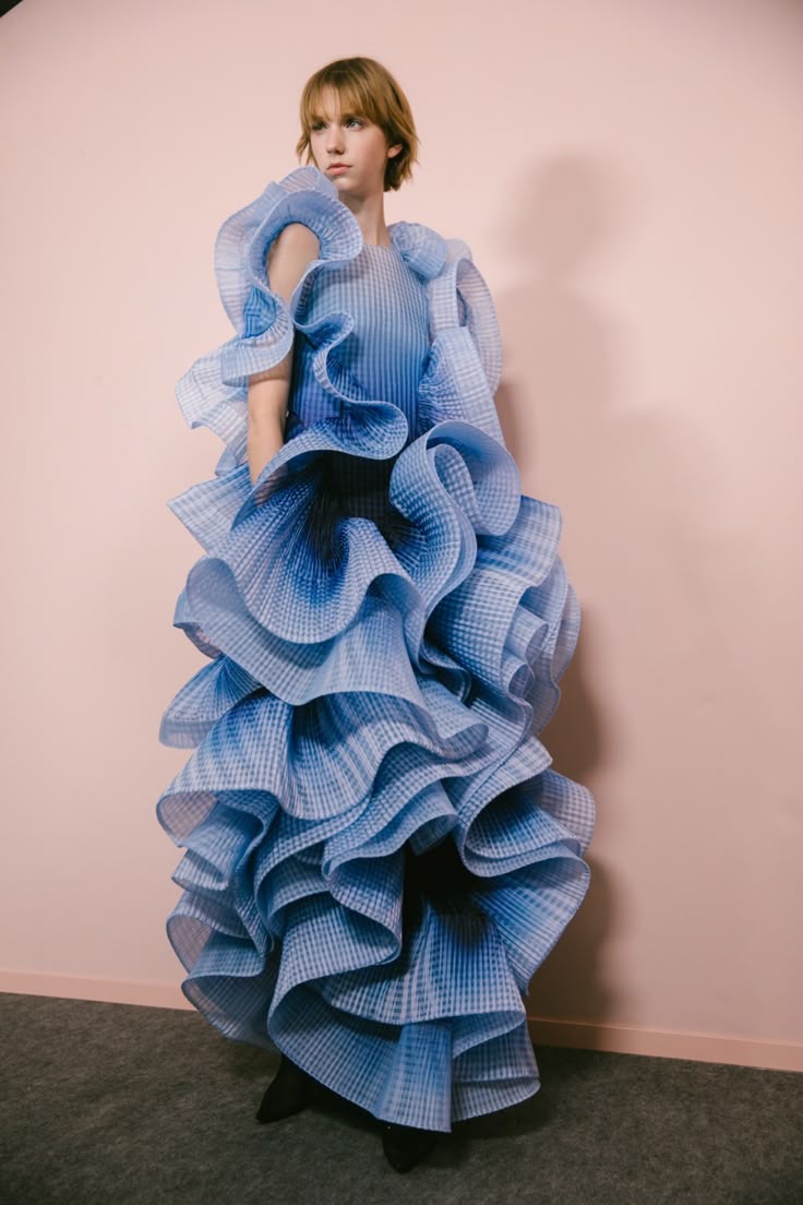 a young, fairskinned model with short blonde hair stands against a light pink wall, wearing a striking blue dress made of many layers of ruffles. the dress has a gradient from light blue to dark blue, and the ruffles create a voluminous, eyecatching effect. the model is looking off to the left, and the overall impression is one of high fashion and artistic design. the floor is a gray carpet or mat.