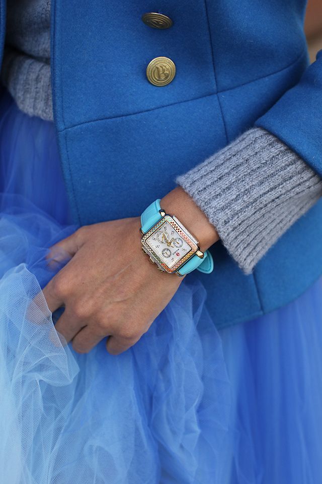 loseup view of a woman wearing a vibrant blue coat and a light blue tulle skirt.  the focus is on her wrist, where she's wearing a turquoise and gold squarefaced watch.  the image showcases the textures of the fabrics and the detail of the watch. the overall style is elegant and fashionable.