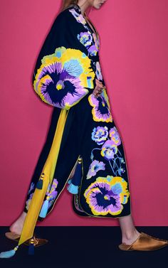 a woman is posing in a long blue floral dress with yellow, pink and purple flowers on it. the dress has long sleeves and a slit on the side. the model is wearing brown sandals and standing against a pink background.