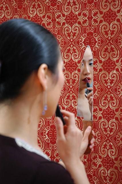 a woman is looking into a mirror, applying lipstick. the mirror is on a wall with a floral pattern and a gold trim. the woman has long black hair, is wearing a black top, and has gold earrings. the background is a red wall with a gold and red ornate floral pattern. the woman is applying lipstick with a red lipstick tube. the woman's reflection is in the mirror and her hair is styled in a ponytail.