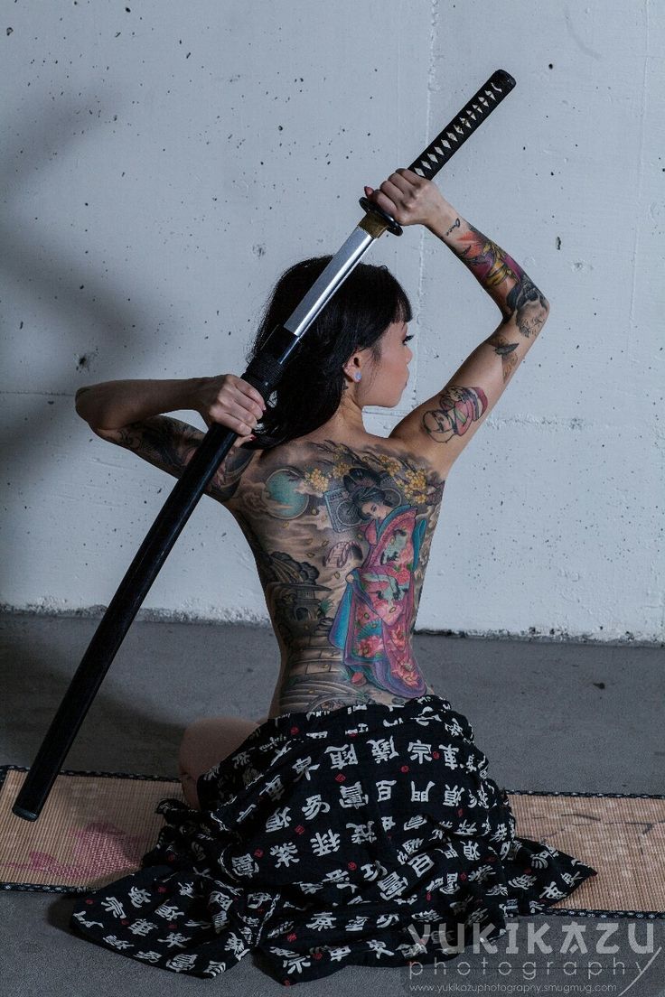 a woman with long black hair and tattoos all over her body is sitting on the floor with a katana sword held above her head. she is wearing a traditional japanese kimono and has an expression of intensity. the background is a concrete wall.