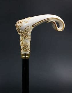 an ornate walking stick with an ivorycolored, carved handle featuring floral designs. the handle has a curved shape and a gold trim. the shaft of the cane is black. the background is a dark grey or black.
