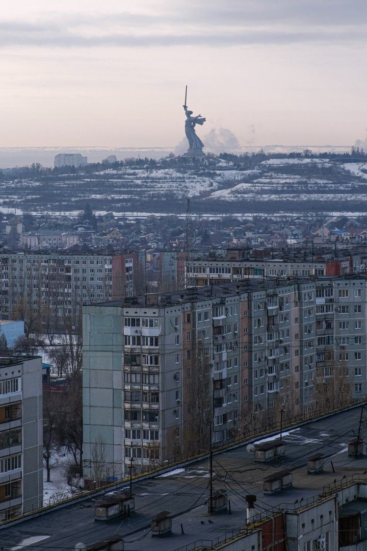 a long shot of the motherland calls statue in volgograd, russia, viewed from a distance across a city of sovietera apartment buildings in the winter. the statue stands prominently on a hill in the background, while the foreground is filled with numerous grey apartment blocks covered in some snow. the sky is overcast and the overall mood is somber and cold.  the image provides a stark contrast between the imposing monument and the everyday life of the city.