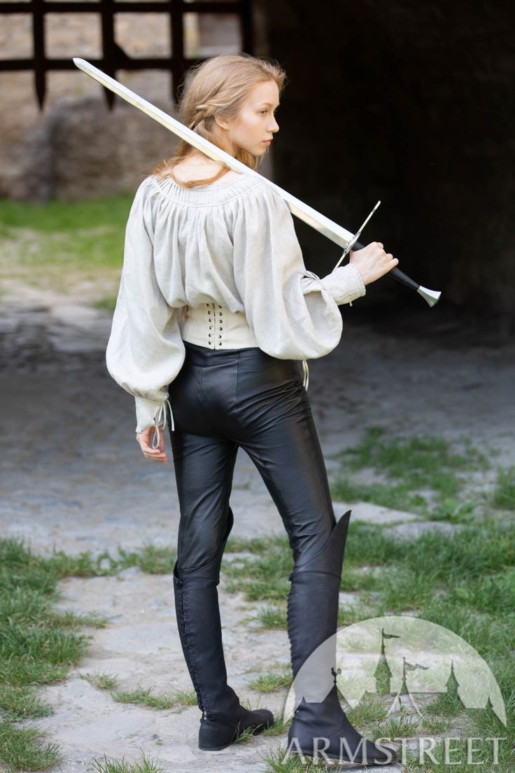 a woman dressed in a medieval costume stands on a path. she is holding a sword across her back and is looking away from the camera. her costume consists of a white blouse, leather pants, and kneehigh leather boots.  she appears to be a warrior.