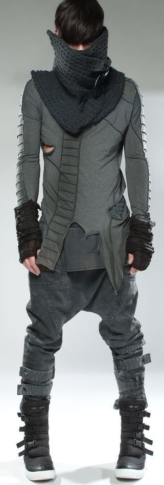 a male model is standing in a studio wearing a black and gray futuristic clothing with an intricately woven scarf, the clothing is styled in a layered fashion with the pants being loose and stylish, the man is also wearing black boots and a black belt. the model is looking slightly down while posing for the photograph. the background is a simple gray.