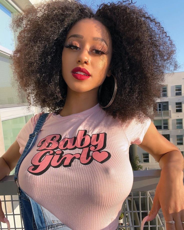 loseup portrait of a young black woman with a large afro hairstyle. she is wearing a pink "baby girl" tshirt and denim overalls. she has on red lipstick and gold hoop earrings. she is posing outdoors on a balcony. the image is bright and sunny.
